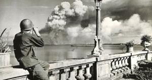 A helmeted US soldier leans on a stone balustrade and looks through binoculars across the bay at smoke rising over Manila in February 1945.
