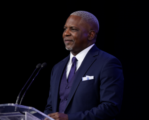 Curtis Caruthers speaking behind a transparent podium during a live presentation, dressed in a navy blue suit, dark vest, white shirt, and tie.