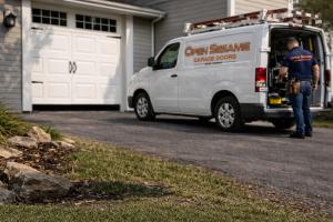 Open Sesame Garage Doors branded service van parked in a driveway while a technician prepares tools for a garage door service.