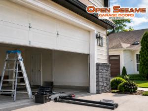 Residential driveway with a ladder, tool box, and replacement torsion springs laid out in front of an open garage door.