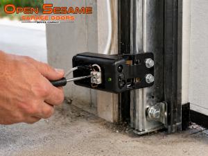 Technician using a screwdriver to adjust and calibrate a black garage door safety sensor with an illuminated green indicator light.