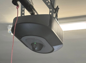 Bottom view of a modern black garage door opener equipped with a built-in security camera lens and indicator status light.