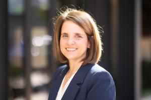 A smiling woman with shoulder-length brown hair wearing a navy blazer and white blouse, photographed outdoors with a softly blurred background.