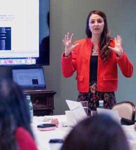 A woman with long dark hair and a red jacket has her hands raised, palm forward, as lectures on her research