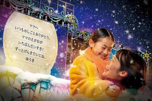 Mother and daughter embracing under falling snow at Universal Studios Japan's Joy of Lights show, with a glowing handwritten message board lit up beside them