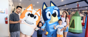 Family poses with Bluey and an orange cartoon dog mascot inside an indoor play center.