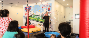 Children and adults watch a man speaking at an indoor climbing-themed event, with a “Clovis Gateway to the Sierras” sign and blue pool.