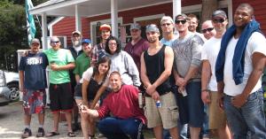 A group of Dismas residents, staff, and volunteers are gathered in front of a red, Vermont-style building with an open flag.