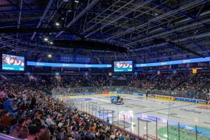 The o2 Belfat is full of hockey fans watching a game. Players are on the ice and around the stadium an LED ribbon screen lines the seats. Two large displays are also visible.