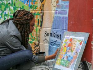 A woman with long braids and glasses sits cross-legged beside a colorful mural reading “Sunkist,” painting a framed illustration of a face with red fruit and leaves propped against a red wall.