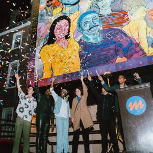 Six people stand on a stage with arms raised as confetti falls at night, beneath a large, colorful mural of portraits in yellow, blue, and purple, with a podium and microphone to the right.