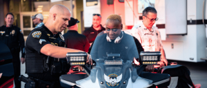 Police officer assists a man seated on a police motorcycle while officers and bystanders gather near an emergency vehicle.