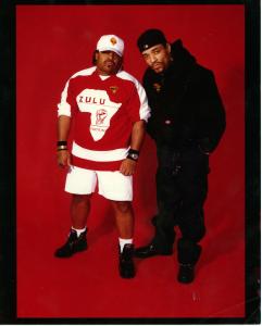 Photograph of hip-hop pioneers Ice T and DJ Afrika Islam standing together against a bright red backdrop. One wears a red-and-white Zulu Nation jersey and white shorts, while the other wears black clothing and a black baseball cap.
