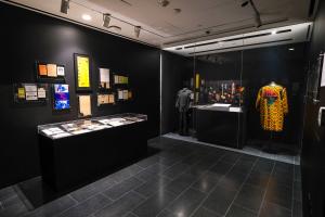 Installation view of DAY ONE DNA at Harvard University's Alain Locke Gallery, featuring archival hip-hop ephemera, framed documents, artifacts in glass display cases, and historic clothing and accessories against black gallery walls.