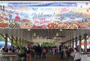 Visitors sit at picnic tables and explore booths beneath a Turkish Food and Culture Day banner with the Wellspring Community Center logo.