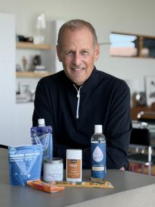 Dave Coulier smiles behind a kitchen counter displaying AwearMarket products, including a white Rustic MAKA Amber Oryn deodorant stick with other beverages, a jar and packaged snacks.