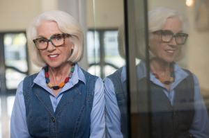 Judy K. Herman smiling beside a glass window that reflects her image, wearing glasses, a blue shirt, and a denim vest.
