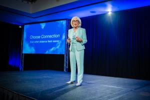 Judy K. Herman speaking on stage in a light blue suit while wearing a headset microphone and holding a presentation remote.