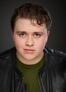 Professional headshot of Cameron Wight wearing a black leather jacket over an olive-green shirt, with curly brown hair and a neutral charcoal-grey background.