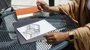A person using the iFLYTEK AINOTE 2 E Ink notebook for digital sketching at an outdoor table, with a stylus and protective case.