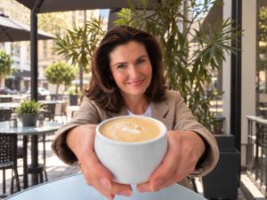 AI-edited image of Linda Cunningham smiling and holding a coffee cup towards the camera at an outdoor cafe.