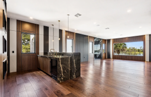 Open room with hardwood floors, dark veined stone bar with brass fixtures, wood wall paneling and windows facing a lake