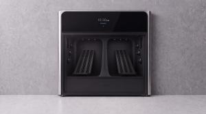 AiRET AX1 shoe care appliance in brushed stainless steel with a black glass door, shown from the front against a gray wall