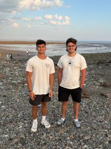 Two boys in white T-shirts and shorts are standing on a rocky shoreline