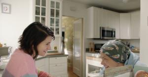 Devin Sidell and Jaime King laughing together in a bright kitchen in a scene from “Love, Danielle,” with Jaime King wearing a patterned headscarf.