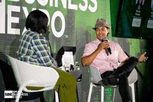 A seated panelist holding a microphone speaks with a moderator against a Small Business Expo stage backdrop.