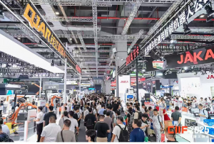 Crowds walk between exhibition stands displaying robots and industrial equipment in a large exhibition hall.