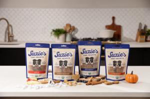 Four stand-up pouches of Suzie's CBD Hearts dog treats in Apple, Peanut Butter, Blueberry, and Pumpkin flavors displayed on a white countertop with fresh ingredients like apples, peanuts, blueberries, and a mini pumpkin.
