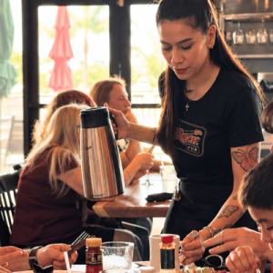 Waitress with tattoos serves coffee from a thermos to diners at a busy restaurant table.