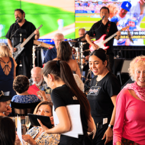 Waitstaff serve guests at a lively concert venue as musicians perform onstage in the background.