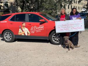 J. Blanton Plumbing team member and a Border Tails Rescue representative stand next to a red J. Blanton Plumbing SUV holding an oversized $5,000 check, with a brindle puppy sitting at their feet.
