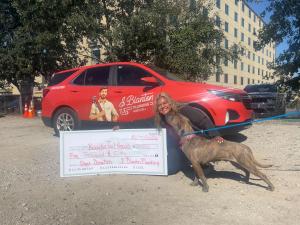 J. Blanton Plumbing team member kneels beside a red J. Blanton Plumbing SUV, smiling as a brindle dog on a leash kisses her face, with an oversized $5,000 donation check propped in front of her.