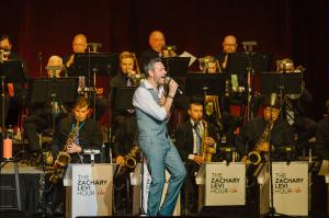 Zachary Levi in a gray suit pant/gray vest outfit singing onstage in front of a 17-piece band and orchestra, with the bandstands reading "Zachary Levi Hour-ish"