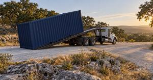 A white tilt-bed truck with its bed tilted, sliding a navy-blue 20ft shipping container onto a gravel pad among live oaks