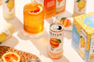 Open can of Oliver Winery Blood Orange Spritz with a glass of spritz, Italian Lemon Spritz cans and 4-packs on a sunny table