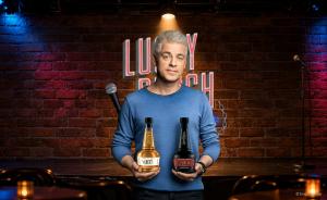 Michael Mittermeier hält die beiden Whiskyflaschen Lucky und Punch von St. Kilian vor einer Backsteinwand mit der Leuchtschrift Lucky Punch