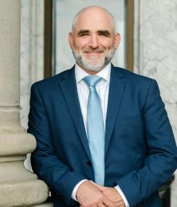 image of trial attorney Kristian Beckett, standing against a courthouse wall wearing a dark blue suit and light blue neck tie.