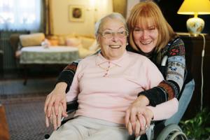 Smiling elderly woman in a wheelchair at home with her adult daughter hugging her from behind
