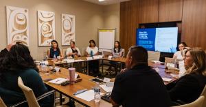 A group of Indigenous men and women sit in a circle around a group of medium-toned wood tables in a board room. Indigenous wood relief art hangs on the wall to the left. On the right wall is a television with a slide on screen saying "About the Indigenous