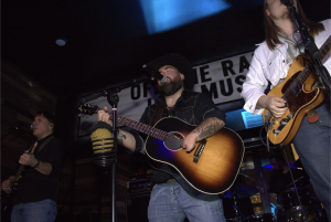 Three musicians perform live on stage at Off The Rails Boston, featuring acoustic and electric guitars under blue stage lighting.