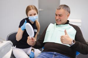 Male dental patient smiling at his restored teeth in a handheld mirror while sitting with his dentist