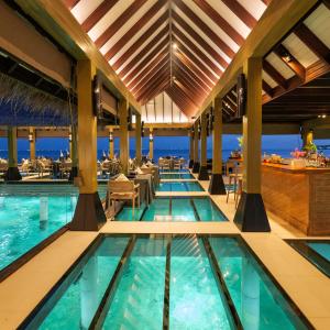 Interior of FireDoor overwater restaurant at Grand Park Kodhipparu Maldives, featuring illuminated glass-floor panels above turquoise lagoon waters, elegant dining tables, a high timber ceiling and ocean views at dusk.