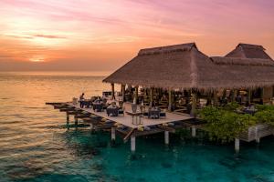 Exterior of FireDoor overwater fine-dining restaurant at Grand Park Kodhipparu Maldives, featuring thatched-roof architecture, outdoor dining terraces above the turquoise lagoon and a pink and orange Indian Ocean sunset.