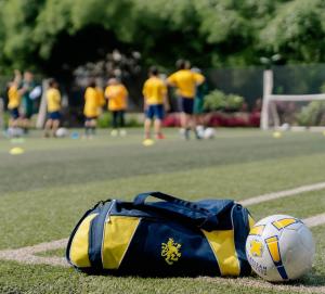 Navy and yellow ISJ sports bag with the gold lion crest beside a football on an artificial pitch, with pupils in yellow shirts training in the background.