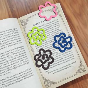 Set of four colorful flower-shaped MDF bookmarks in pink, green, blue, and brown placed on the pages of an open book on a wooden table.