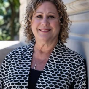 Alt Text: Debbie Driskell, Delaware Township Trustee in Hamilton County, Indiana, smiles warmly in an outdoor portrait. She has shoulder-length curly blonde hair and wears a black-and-white patterned blazer over a black top, with a silver necklace.